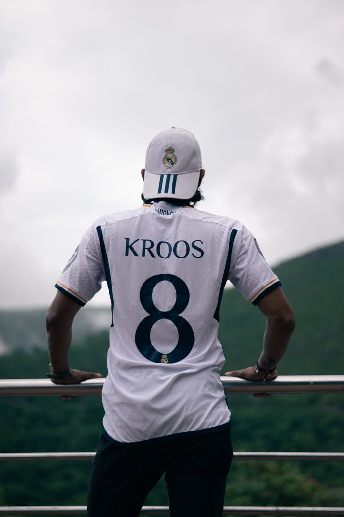Man in a Kroos jersey leans on railing, enjoying an outdoor view.