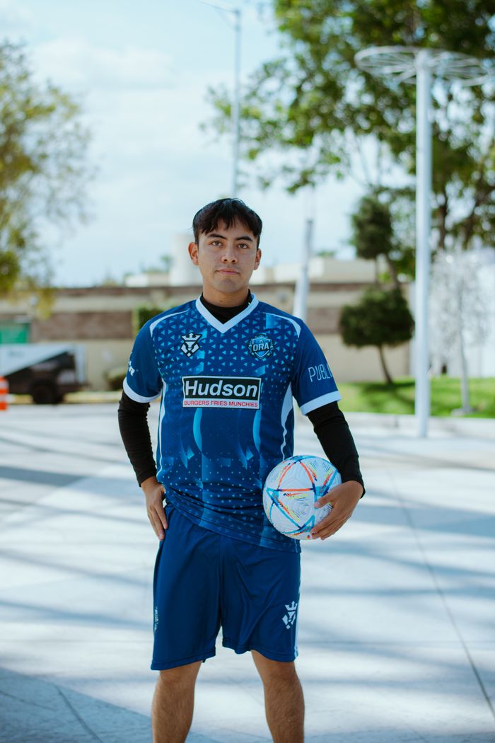 Young male footballer poses outdoors wearing blue jersey with soccer ball in hand.